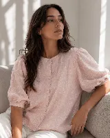 Blusa de verano en color rosado con acabado brillante, de corte estructural y silueta holgada. Presenta cuello redondo, cierre frontal con botones de nácar ocultos por una cartera y mangas cortas abullonadas con tablas que aportan volumen.