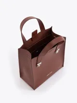 Cartera tipo tote pequeña de cuero color tostado, con doble asa superior y herrajes metálicos plateados en forma de llama en la base de las manijas. Presenta el logo de la marca estampado en la parte inferior frontal.