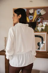 Blusa blanca de manga larga con cuello redondo estilo bebé, cierre frontal con botones y detalle de volados en puños y cintura.