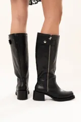 Bota estilo biker de cuero negro con caña alta y cierre metálico lateral. Presenta un diseño versátil con estribo y cadena desmontables, taco cuadrado y suela con plataforma.