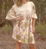 Vestido corto acampanado color crudo, con estampado artesanal de hojas en tonos marrones, violetas y amarillos. Tiene cuello redondo y mangas cortas amplias.