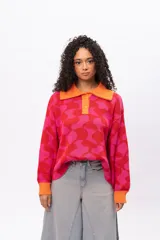 Buzo de tejido de punto con diseño geométrico en tonos fucsia y rojo, cuello tipo polo con cierre de botones y puños en color naranja contrastante. Presenta un corte holgado y oversize.