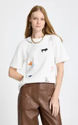 Remera blanca de algodón con cuello redondo y mangas cortas. Presenta un estampado disperso de perros de diferentes razas y colores, junto con un detalle de texto bordado cerca del cuello.