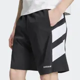 Short Adidas Adicolor Double Blocked de color negro, confeccionado en tejido ripstop. Presenta un diseño holgado, bolsillos laterales, cierre de cremallera, el logo del Trifolio bordado y detalles gráficos de franjas blancas en el lateral.