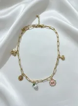 Collar dorado con cadena de eslabones rectangulares y varios dijes: trébol, moneda, corazón, fresa y medalla rosa con la letra M.