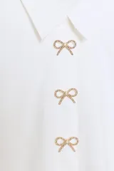 Camisa de popelín blanca con cuello solapa y manga larga. Presenta detalle de botones joya decorativos en forma de lazo en la parte frontal y cierre oculto por solapa.
