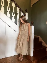 Vestido midi sin mangas de lino color beige, con cuello camisero, botonadura frontal, volados decorativos en el pecho y un lazo para atar en la cintura.