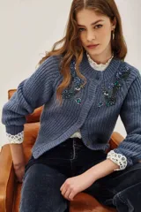 Cardigan de punto acanalado color azul grisáceo, de corte cropped y abotonadura frontal. Presenta apliques florales bordados con cuentas y lentejuelas en tonos azules y verdes en la parte delantera.