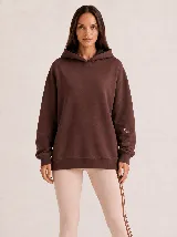 Hoodie Trena de frisa color marrón chocolate, con moldería oversize, hombros caídos, capucha con cordones ajustables y bolsillo tipo canguro. Presenta puños y ruedo de rib elástico, y detalles de bordado en la manga.