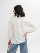 Camisa blanca de algodón con delicados bordados florales y líneas verticales sutiles, cuello camisero clásico y cierre frontal de botones.