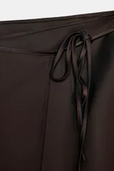 Falda corta tipo pareo, color marrón oscuro, confeccionada en tejido satinado con lazada ajustable en el lateral.