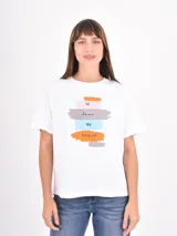 Remera blanca de algodón con cuello redondo y mangas cortas. Presenta un estampado frontal con pinceladas de colores y la frase 'in love we trust'.