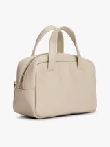 Cartera de mano tipo bowling de Tommy Jeans, color beige, con doble asa superior y correa ajustable extraíble. Presenta un diseño estructurado con cierre superior y logo metálico de la marca en la parte frontal.