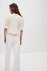 Blusa color blanco crudo, de escote en V pronunciado con ribetes de volado, mangas abullonadas al codo y detalles de alforzas, encaje y bordados en el frente.