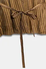 Camisa marrón a rayas con cuello redondo, mangas largas anchas y puños rectos. Cuenta con cierre frontal de botones y un lazo cinturón ajustable del mismo tejido en la cintura.