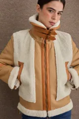 Campera tipo gamulán de cuero color camel con interior y detalles de piel sintética en color crema. Presenta cuello alto con doble hebilla ajustable, cierre frontal con cremallera y bolsillos laterales con cierre.