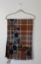 Bufanda de gran tamaño con diseño de cuadros en tonos naranja, beige y azul, decorada con un bordado floral en color azul oscuro y terminaciones con flecos.