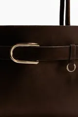 Bolso tote de acabado suave, color marrón oscuro, con asas tubulares y detalle de correa frontal con hebilla metálica dorada.
