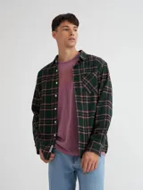 Camisa de manga larga a cuadros azules, confeccionada en viyela 100% algodón, con bolsillo en el pecho y botones de pasta. Presenta una etiqueta de cuero sintético en el ruedo y otra en la cartera.