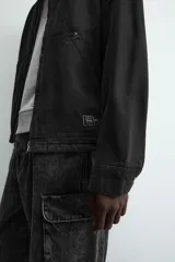 Cazadora de algodón color negro lavado, de corte relajado (relaxed fit), con cuello solapa a contraste, cierre frontal de cremallera, bolsillos canguro en la cadera y puños con botón a presión.