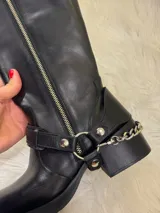 Bota alta tipo biker de cuero negro con cierre metálico lateral que recorre toda la caña. Presenta un diseño versátil con estribo y cadena metálica desmontables en la zona del tobillo, taco cuadrado y plataforma delantera.