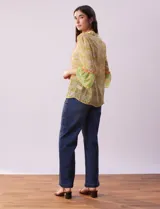 Blusa de gasa estampada con flores amarillas y verdes, marca Alex & Lili. Tiene cuello mao, mangas 3/4 acampanadas y cierre frontal con botones. Presenta volados verticales color naranja en el frente y en las mangas.