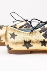 Zapatos tipo blucher artesanales de napa dorada con estampado de estrellas negras. Presentan un adorno de flecos de napa negra en el empeine, cierre con cordones y suela de cuero.
