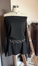 Vestido negro de corte holgado con escote tipo bote y mangas largas. Presenta un diseño minimalista con cintura elástica y se complementa con un cinturón de cadena metálica con eslabones circulares.