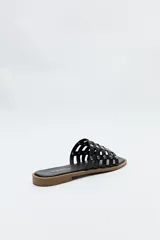 Sandalia baja negra, tipo mule o chinela, con capellada de tiras entrelazadas y apliques metálicos redondos (tachas). La suela es plana y de color marrón claro.