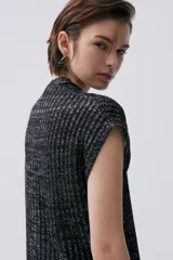 Kimono tejido de punto en color crudo con sutil brillo de lurex. Presenta un diseño sin mangas, corte largo y cierre frontal mediante dos lazos finos. El tejido tiene una textura acanalada vertical.