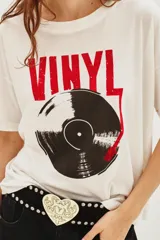 Remera de algodón blanca, corte holgado, con cuello redondo. Presenta un estampado frontal grande con la palabra "VINYL" en rojo y la ilustración de un disco de vinilo en negro y rojo.