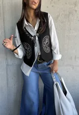 Chaleco negro de gamuza con bordados blancos estilo paisley y cierre frontal con cordones y detalles metálicos.