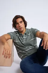 Remera tipo polo de manga corta en color verde oliva, con diseño de rayas horizontales en tonos blanco y azul marino. Presenta cuello tejido acanalado, cierre frontal con botones y logo bordado en el pecho.