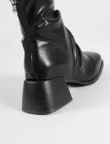 Botas de caña media en color negro, confeccionadas en cuero sintético con acabado ligeramente arrugado. Presentan una puntera cuadrada, cierre posterior y un taco cuadrado de altura media con textura estriada.