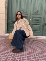 Campera tipo bomber de gamuza color beige, con diseño bordado de flores y motivos geométricos en blanco y negro. Presenta cuello clásico, cierre frontal con botones a presión y puños y ruedo de tejido elástico acanalado.