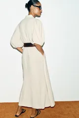 Vestido midi color crudo, con cuello camisero, mangas abullonadas por debajo del codo con puños elásticos y cierre frontal oculto con botones. Incluye bolsillos laterales y un cinturón ancho de cuero marrón oscuro con hebilla metálica.