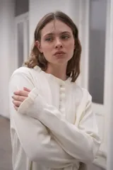 Sweater de punto color crema, con cuello redondo y cierre frontal parcial con botones. Presenta mangas largas con puños acanalados y detalle de volados en los extremos.