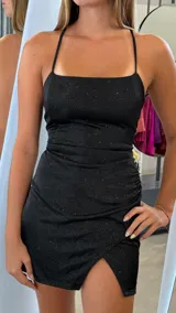 Vestido corto negro con brillo, corte ajustado y escote cuadrado con tirantes finos cruzados en la espalda. La falda presenta un cruce frontal con frunce lateral.