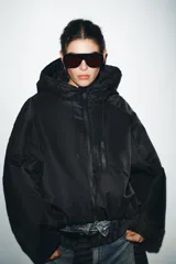 Chaqueta acolchada de color negro con acabado repelente al agua y resistente al viento. Presenta cuello subido con capucha ajustable, cierre frontal con cremallera termosellada, bolsillos delanteros con cremallera y bajo con ajuste elástico. Las mangas largas cuentan con tiras adhesivas ajustables en los puños.