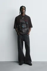 Camiseta negra de algodón con corte boxy fit y cuello redondo. Presenta un estampado gráfico desgastado con temática espacial o celestial en tonos azules y blancos, y texto en la parte superior e inferior.