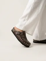 Zueco de cuero color marrón con diseño artesanal, presenta una hebilla metálica decorativa, tachas y remaches plateados, además de detalles de costuras bordadas en el empeine.