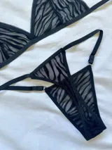 Conjunto de lencería de dos piezas confeccionado en tul transparente con estampado animal print tipo cebra aterciopelado en color negro. Incluye un corpiño triangular sin aro con breteles regulables y una bombacha tipo tanga a juego con detalles de argollas metálicas en los laterales.