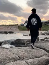 Canguro negro con capucha y cordones ajustables. Estampa en la espalda con el texto "Chema Surf Trip" y dibujos de un sol, una tabla de surf, una carpa y otros elementos relacionados con el surf.