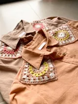 Camisa tostada de manga larga con cuello clásico decorado con apliques de crochet en tonos marrones, blancos y verdes, cierre frontal de botones y diseño holgado.