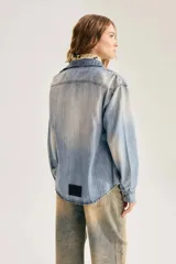 Camisa de denim azul con lavado gastado, bolsillos con solapa en el pecho, cuello en punta y cierre frontal de broches metálicos.