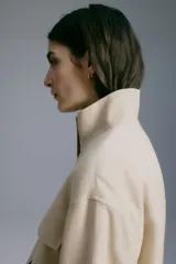 Campera beige con cuello chimenea, botones a presión ocultos en la parte delantera, bolsillos superiores con solapa y bolsillos laterales. Cuenta con diseño forrado y ajuste con cordón elástico en el bajo.