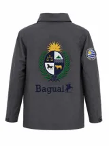 Campera impermeable de tela taslon color gris oscuro, con diseño de cuello alto y cierre frontal. Presenta un gran bordado en la espalda con el escudo nacional uruguayo y el logo de la marca Bagual, además de un parche circular con la bandera de Uruguay en la manga.