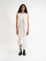 Vestido midi blanco sin mangas, con cuello redondo y diseño holgado. Presenta un cordón ajustable fruncido en el centro de la parte frontal y parches circulares sutiles en la tela.