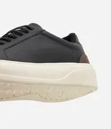 Championes casuales para hombre confeccionados en cuero negro, con diseño minimalista de caña baja, detalles en color marrón en el talón, cordones elásticos y suela alta de goma en tono off-white.