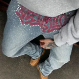 Pantalón de jean con corte recto y acabado desgastado, decorado con pedrería brillante en toda la superficie y un diseño de graffiti en color rojo con cristales en la zona frontal.